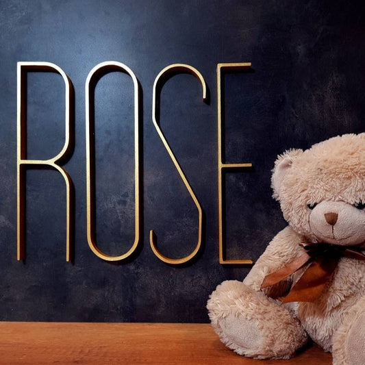 Gold skinny letters ROSE Mid century modern minimalistic name sign on a black backdrop next to a teddy bear on a shelf in a nursery