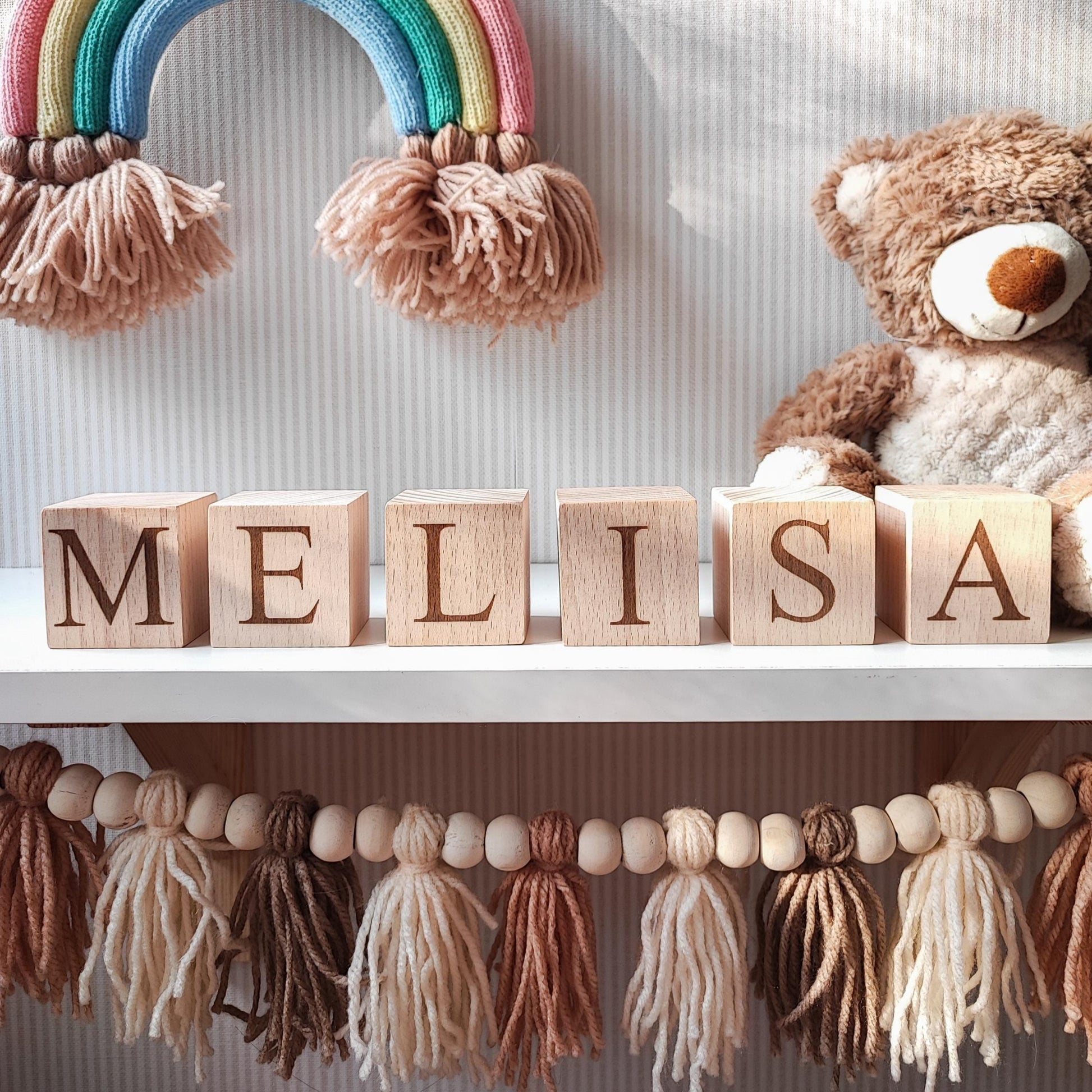 MELISA name engraved on natural wooden blocks, standing on a white nursery shelf next to boho baby decorations