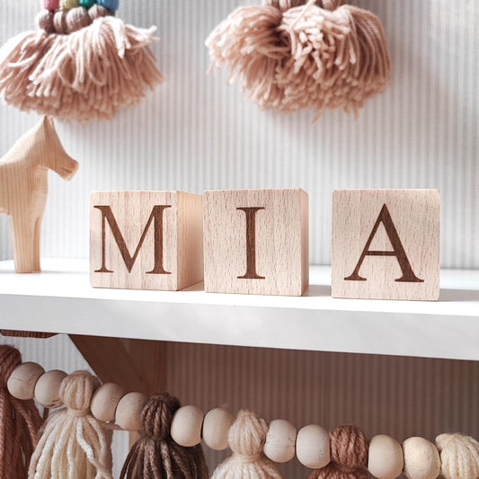 MIA name engraved on natural wooden blocks, standing on a white nursery shelf next to boho baby decorations